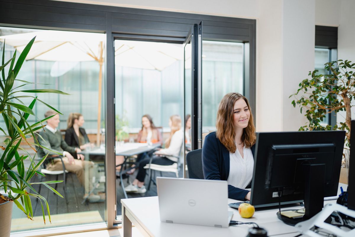 Mitarbeiterin von AMW im Büro, im Hintergrund mehrere Mitarbeiterim Gespräch auf der Terrasse